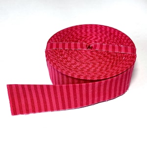 May include: A spool of fabric ribbon featuring a vibrant pattern of alternating vertical stripes in two shades of red. The ribbon is tightly wound into a circular coil, with one end extending outward to display the consistent striped design.