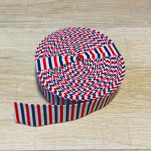 May include: A roll of red, white, and blue striped fabric ribbon. The ribbon is rolled up and the end of the ribbon is laying flat.