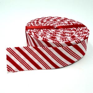 May include: A roll of fabric ribbon featuring a diagonal candy cane stripe pattern in red and white. The ribbon is tightly wound into a circular spool and secured with a thin red satin band wrapped around the center of the roll.