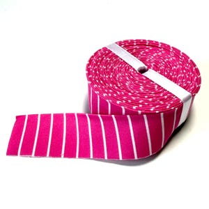May include: A roll of hot pink elastic ribbon featuring vertical white stripes. The fabric band is wound into a tight circular spool and secured with a white paper strip across the top, displaying a consistent striped pattern throughout.
