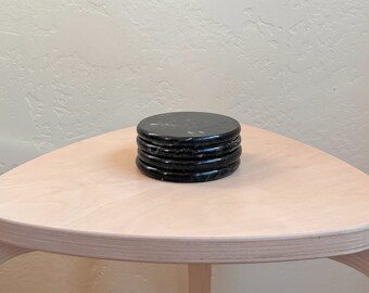 May include: A stack of four circular black marble coasters with subtle white veining. Each coaster has a smooth, polished finish and a consistent round shape, neatly piled on top of one another to form a single vertical column.