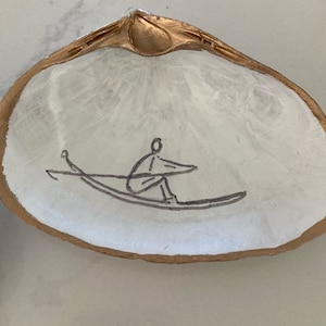 Może przedstawiać: Biała muszla z pomalowaną na złoto krawędzią. Prosty czarny rysunek liniowy przedstawiający osobę wiosłującą w łodzi znajduje się na środku muszli. Muszla leży na białej powierzchni.