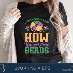 May include: A black t-shirt with a Mardi Gras theme design. The design features a purple, green, and gold fleur-de-lis, beads, and the text "Does your mama know how you got those beads?"