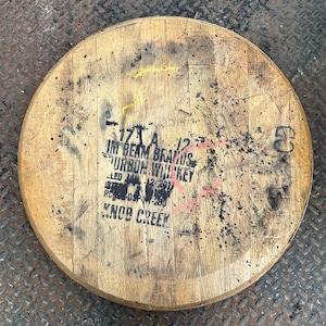 May include: Circular wooden barrel lid with black text and markings. The text reads "Jim Beam Brands Bourbon Whiskey" and "Knob Creek". The wood has a natural, aged appearance with dark spots and yellow paint marks.