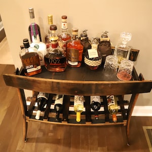 May include: A rustic wooden bar cart with a dark top, holding various bottles of liquor and wine. The cart has a lower rack for wine bottles and is set on a hardwood floor. Several glasses are also on the cart.