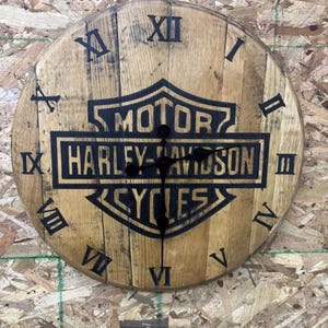 May include: A round wooden clock with the Harley-Davidson logo in black. The clock face is made of wood and features black Roman numerals and hands. The clock is mounted on a textured wooden surface.