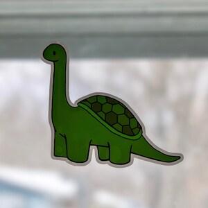 May include: A green dinosaur sticker with a long neck and a turtle-like shell. The sticker has a white outline and is likely made of vinyl. The dinosaur is a cartoon style and has a friendly expression.