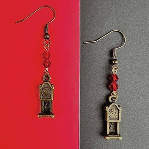 Puede incluir: Pendientes colgantes con un dije de reloj antiguo en miniatura. Los pendientes tienen un acabado dorado, con tres cuentas facetadas rojas. El dije del reloj tiene una esfera y un péndulo detallados. Fondo rojo y gris.