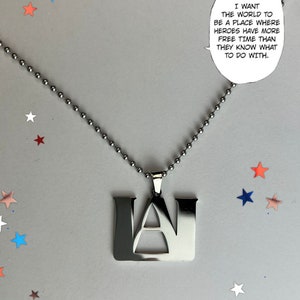 Puede incluir: Un collar con colgante plateado con las letras "UA" en una fuente audaz y cuadrada. El collar cuelga de una cadena de bolas. La imagen incluye una burbuja de diálogo con el texto: "I WANT THE WORLD TO BE A PLACE WHERE HEROES HAVE MORE FREE TIME THAN THEY KNOW WHAT TO DO WITH."
