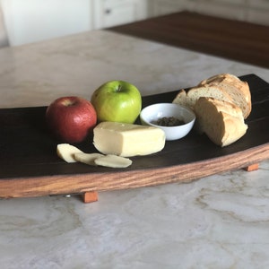 May include: A wooden serving tray made from a reclaimed wine barrel with a dark brown finish. The tray has a curved shape and is supported by three wooden feet. The tray is filled with a variety of snacks, including apples, cheese, bread, and herbs.