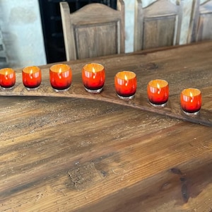 May include: A wooden candle holder with a curved shape, holding seven red and orange glass candle holders. The candle holders are arranged in a row on the holder.