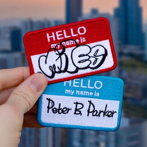以下が含まれることがあります： 刺繍された名前タグが2つ。 上のタグは赤で、白い文字で「HELLO my name is」と書いてあり、黒と白のグラフィックで「WIB」という言葉が書かれています。 下のタグは青で、白い文字で「HELLO my name is Peter B. Parker」と書いてあります。