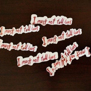 Può includere: Una collezione di adesivi bianchi con la frase "I must not tell lies" scritta in rosso, con un carattere scritto a mano. Gli adesivi sono sparsi su una superficie marrone scuro.