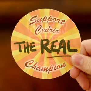 Puede incluir: Una pegatina redonda con un dise&ntilde;o de rayos de sol amarillo y naranja. La pegatina dice "Support Cedric The Real Champion".