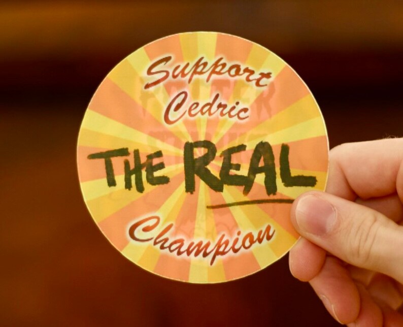 Puede incluir: Una pegatina redonda con un dise&ntilde;o de rayos de sol amarillo y naranja. La pegatina dice "Support Cedric The Real Champion".