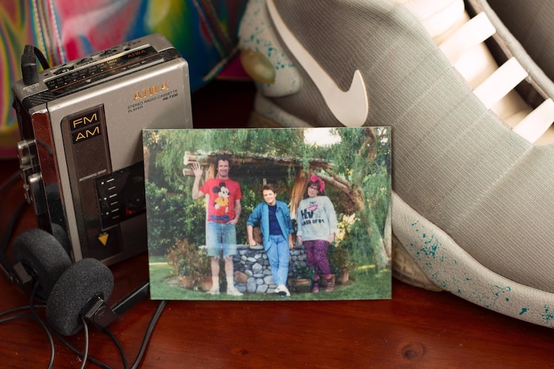 May include: A vintage photograph of three people standing in front of a stone structure. A silver cassette player and headphones are on the left. A gray and white Nike shoe is on the right.