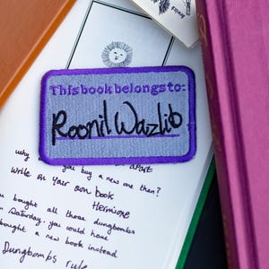 Peut inclure: Une étiquette de livre violette et grise avec le texte "This book belongs to: Roonil Wazlib" écrit en cursive noire.