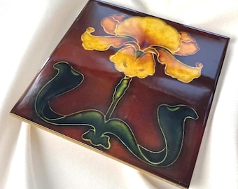 Puede incluir: Un azulejo de cerámica cuadrado con acabado brillante que presenta una flor de iris amarilla central con hojas de color verde oscuro sobre un fondo de color marrón rojizo intenso. El azulejo tiene una superficie lisa y reflectante.