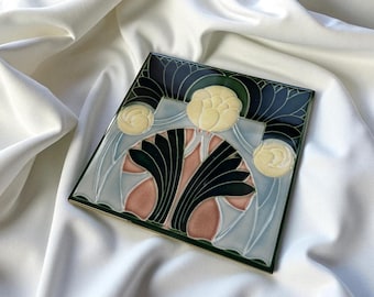 Puede incluir: Un azulejo de cerámica cuadrado con un diseño floral estilizado que presenta tres flores circulares de color crema. El patrón incluye secciones en verde oscuro, azul claro y rosa suave separadas por bordes negros en relieve.
