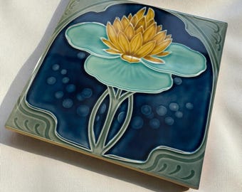 Puede incluir: Un azulejo de cerámica cuadrado con un diseño en relieve de un nenúfar amarillo con pétalos de color verde claro centrado sobre un fondo azul profundo. El borde está decorado con patrones de remolinos verde claro y acentos circulares.
