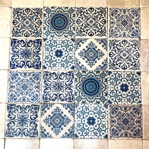 May include: A collection of sixteen square ceramic tiles arranged in a four-by-four grid, featuring a variety of intricate blue and white geometric and floral patterns, surrounded by a border of plain, light beige, marble-textured square tiles.