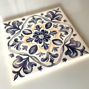 May include: A square ceramic tile featuring a symmetrical pattern in shades of dark blue and muted yellow on a white background. The design consists of intricate floral and leaf-like motifs arranged in a central star shape with corner accents.