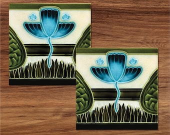 May include: Two square ceramic tiles with a blue flower design. The flower has three petals and is surrounded by green leaves. The tiles have a white background with black and green accents.
