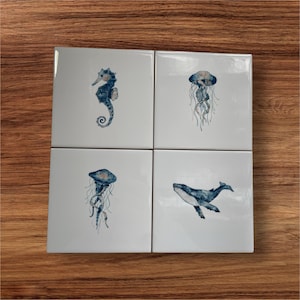 May include: Four white square tiles featuring watercolour illustrations of marine creatures. The images depict a seahorse, two jellyfish, and a whale, in shades of blue and grey. The tiles are arranged on a wooden surface.