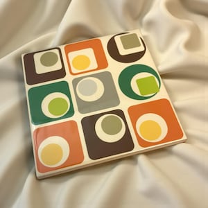 May include: Square ceramic coaster with a repeating pattern of colourful squares and circles. The design features orange, green, brown, and grey squares with contrasting circles in the centre. The coaster has a glossy finish.