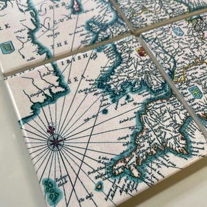 May include: A set of square ceramic coasters with a vintage map design. The coasters have a cream background with teal and brown map details, including text and compass lines. These coasters are perfect for protecting tables from drink marks.