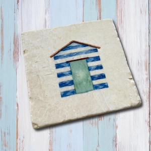 May include: Square coaster with a beach hut design. The hut features blue and white striped walls, a brown roof, and a green door. The coaster is off-white with a textured surface, set against painted wooden planks.