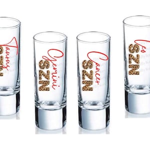 May include: Four clear shot glasses with silver bases. Each glass features a different zodiac sign name in red script, with the letters "SZN" in a leopard print design. The signs are Taurus, Gemini, Cancer, and Leo.