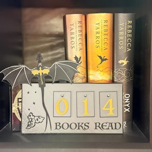 May include: A bookshelf display featuring three books by Rebecca Yarros, with a dragon-shaped bookend and a counter that reads "014 BOOKS READ". The books have gold and black covers.