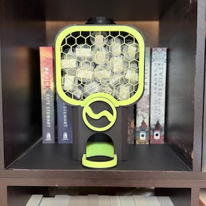May include: A black and neon yellow dispenser featuring a hexagonal honeycomb grid front panel. The unit contains multiple small, clear cylindrical containers filled with white items. A circular logo with a wavy line is centered on the front.