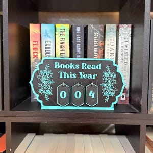 May include: A black rectangular sign with a decorative teal border and floral accents, featuring the text "Books Read This Year" in teal. Three small, hanging black tags with teal numbers "0", "0", and "4" are attached to the bottom.
