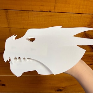 May include: A white dragon mask with sharp teeth and pointed horns. The mask is made of a thick, opaque material and is held up by a hand. The mask is a fantasy-themed item.
