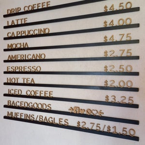May include: A black and gold sign with a menu of coffee drinks and prices. The drinks listed are drip coffee, latte, cappuccino, mocha, americano, espresso, hot tea, iced coffee, and baked goods. The prices range from $2.00 to $4.75. The sign also includes a section for muffins and bagels with prices of $2.75 and $1.50.