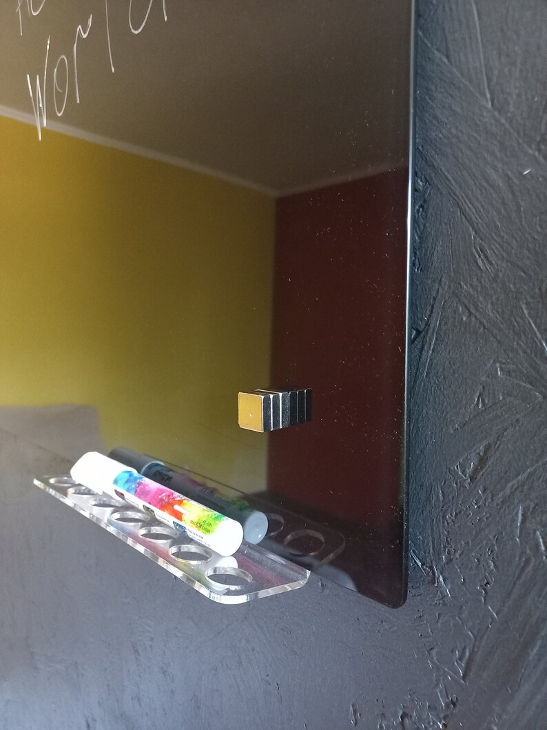 May include: A black glass dry-erase board with the words "the world" written in white. A clear acrylic marker holder with colorful markers is attached to the board. A magnetic holder is also attached to the board.