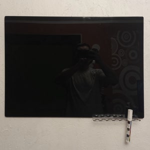 May include: A large, rectangular black dry-erase board mounted on a textured white wall. A clear plastic marker holder with several markers is attached below the board. The reflection of a person holding a camera is visible on the board's surface.