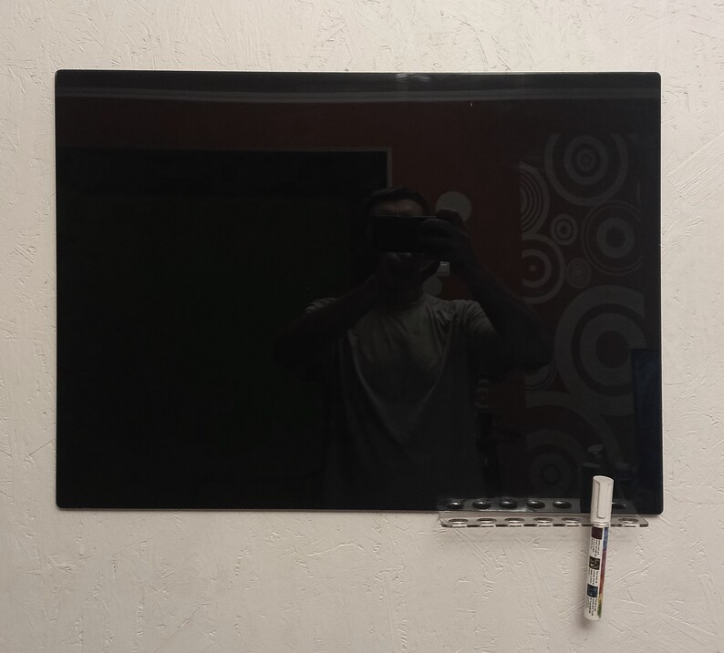 May include: A large, rectangular black dry-erase board mounted on a textured white wall. A clear plastic marker holder with several markers is attached below the board. The reflection of a person holding a camera is visible on the board's surface.