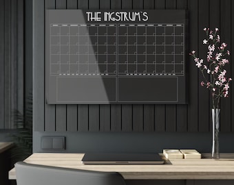 May include: A black dry erase calendar with the text "THE INSTRUMS" at the top. The calendar is divided into two columns, each with a grid of squares for each day of the month. The calendar is hanging on a black wall.