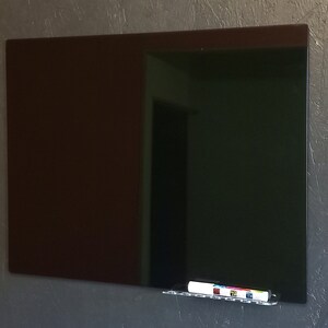 May include: A large, rectangular, dark brown and black glass dry-erase board mounted on a textured gray wall. A clear acrylic shelf holds a white dry-erase marker with colorful accents. The board reflects the room's interior.
