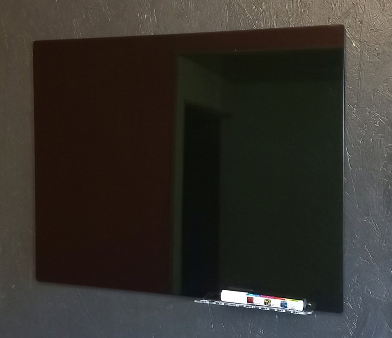 May include: A large, rectangular, dark brown and black glass dry-erase board mounted on a textured gray wall. A clear acrylic shelf holds a white dry-erase marker with colorful accents. The board reflects the room's interior.