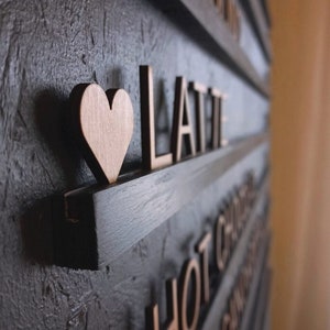 May include: A black wooden sign with the word "LATTE" in gold letters and a small wooden heart. The sign is mounted on a wall with other signs that say "HOT CHOCOLATE" and "APPLE PIE".