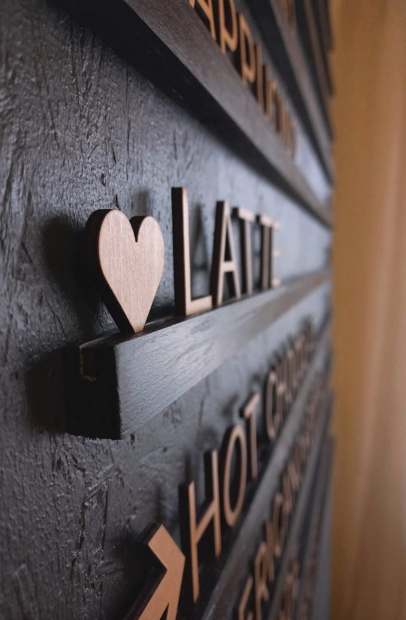 May include: A black wooden sign with the word "LATTE" in gold letters and a small wooden heart. The sign is mounted on a wall with other signs that say "HOT CHOCOLATE" and "APPLE PIE".