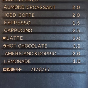May include: A black chalkboard menu with gold lettering lists coffee and pastry items with prices. The menu includes items like almond croissant, iced coffee, espresso, cappuccino, latte, hot chocolate, Americano & Doppio, and lemonade. The prices range from $1.00 to $3.50. The menu also includes social media icons and payment methods accepted.