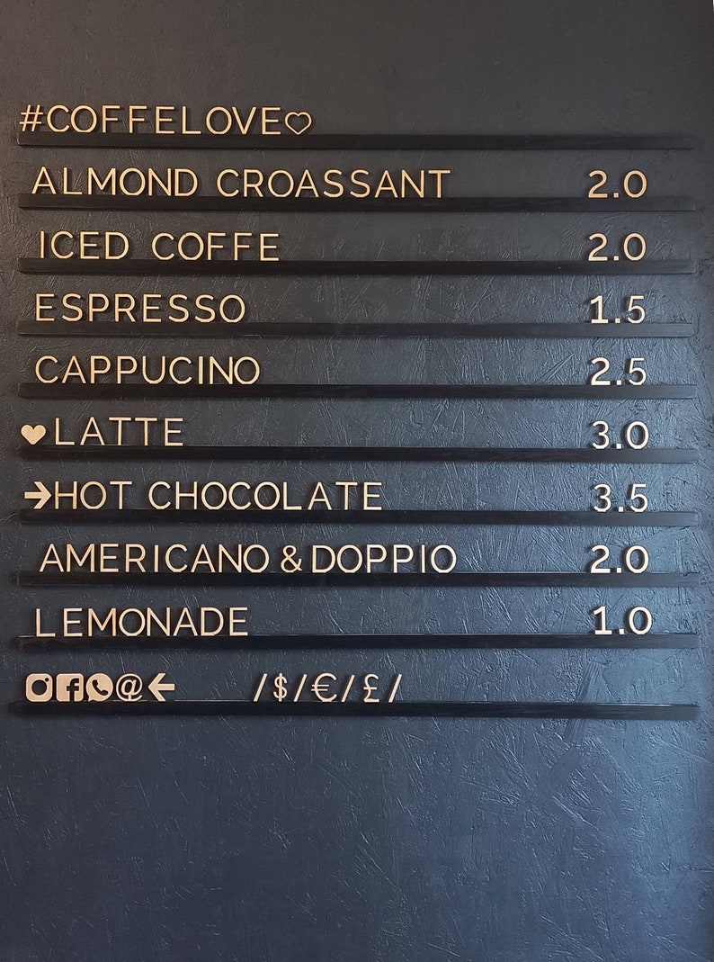 May include: A black chalkboard menu with gold lettering lists coffee and pastry items with prices. The menu includes items like almond croissant, iced coffee, espresso, cappuccino, latte, hot chocolate, Americano & Doppio, and lemonade. The prices range from $1.00 to $3.50. The menu also includes social media icons and payment methods accepted.