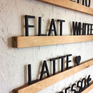 May include: Wooden sign with black letters spelling out different coffee drinks, including "ristretto", "flat white", "latte", and "espresso".