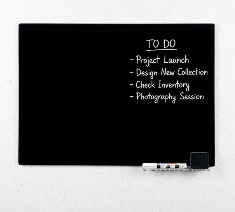 May include: A black rectangular whiteboard with the words "TO DO" and a list of tasks in white. The tasks include "Project Launch", "Design New Collection", "Check Inventory", and "Photography Session". A small eraser and marker holder are at the bottom.
