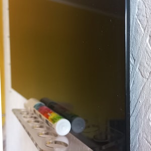 May include: A clear acrylic shelf with several round holes, holding two cylindrical markers. One marker has a colorful label, the other is dark. A large black surface is behind the shelf, reflecting the markers. The shelf is mounted on a white wall.
